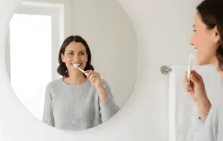Smiling adult brushing teeth in a bright, sunlit bathroom with clean white tones and space for text.