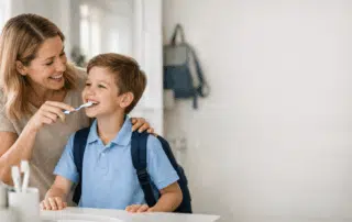 Parent helps a school-aged child brush their teeth in a bright morning bathroom before school.