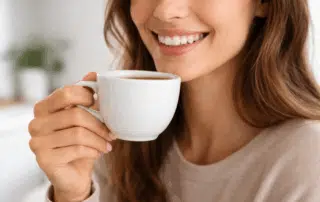 Close-up of a smiling person holding a white coffee cup near their mouth, showing clean, natural-looking teeth in soft natural light.