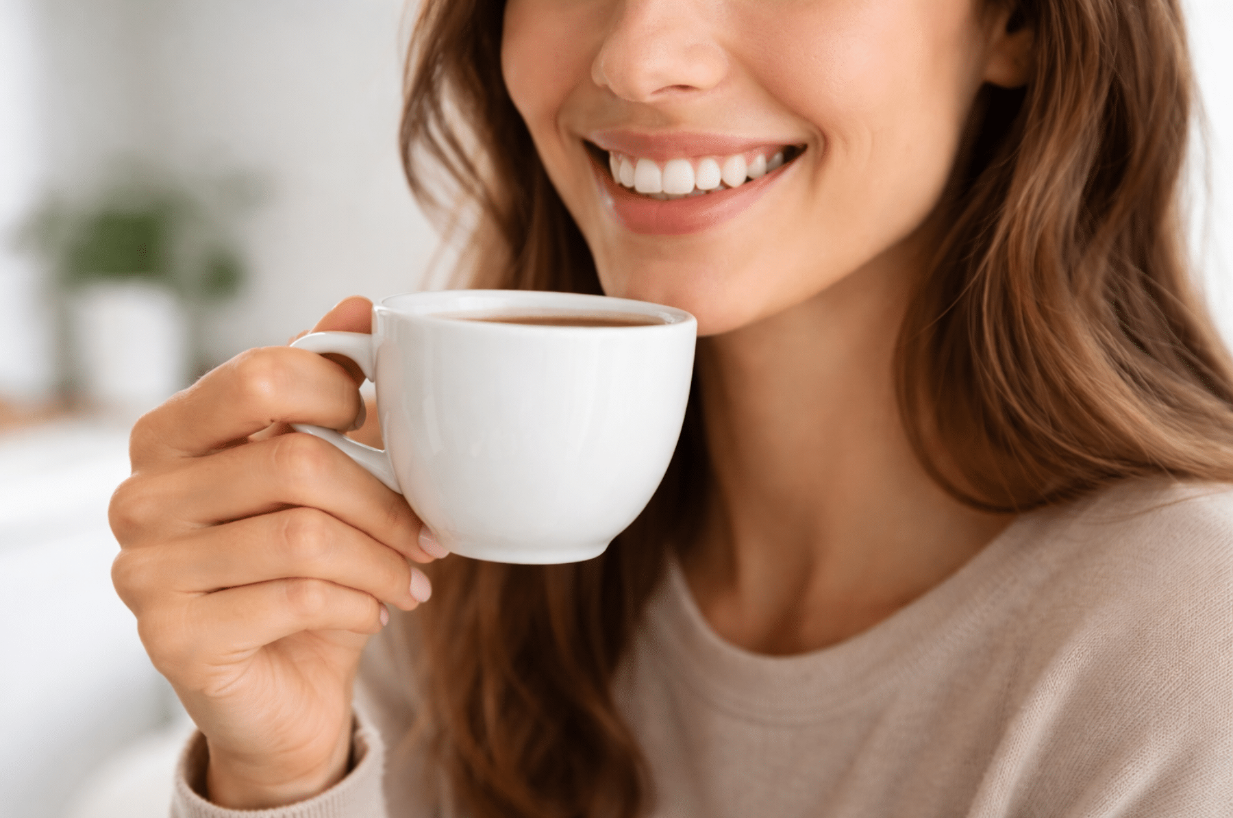 Close-up of a smiling person holding a white coffee cup near their mouth, showing clean, natural-looking teeth in soft natural light.