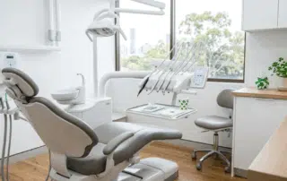 A bright dental treatment room with a reclining chair, overhead light, instruments, and a window view of trees and a city skyline.