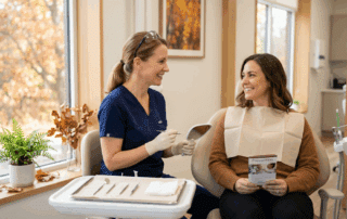 A dentist in scrubs smiles while holding a mirror and tool, speaking with a seated patient wearing a dental bib and holding a pamphlet in a bright, cozy exam room.